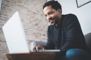 Happy african man dressed in grey pullover and typing on laptop, smiling while sitting the sofa.