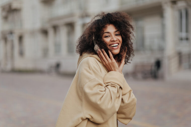 Happy dark-skinned woman in beige hoodie listens to music in headphones outdoors. Curly lady smiles sincerely.