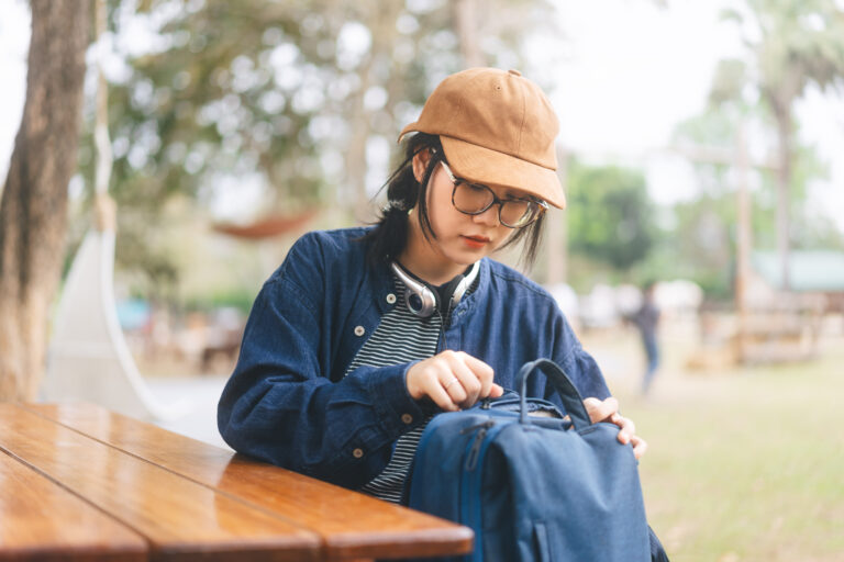 Asian woman sitting on outdoor table open bag. Wearing denim and hat with eyeglasses. Camping area blur background.