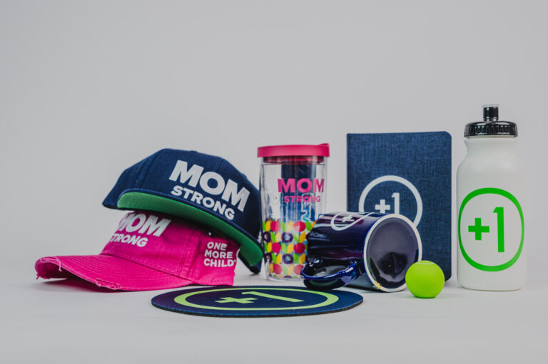 A collection of corporate swag including hats, coffee mug, water tumber, and water bottle, and a notebook for the nonprofit One More Child.