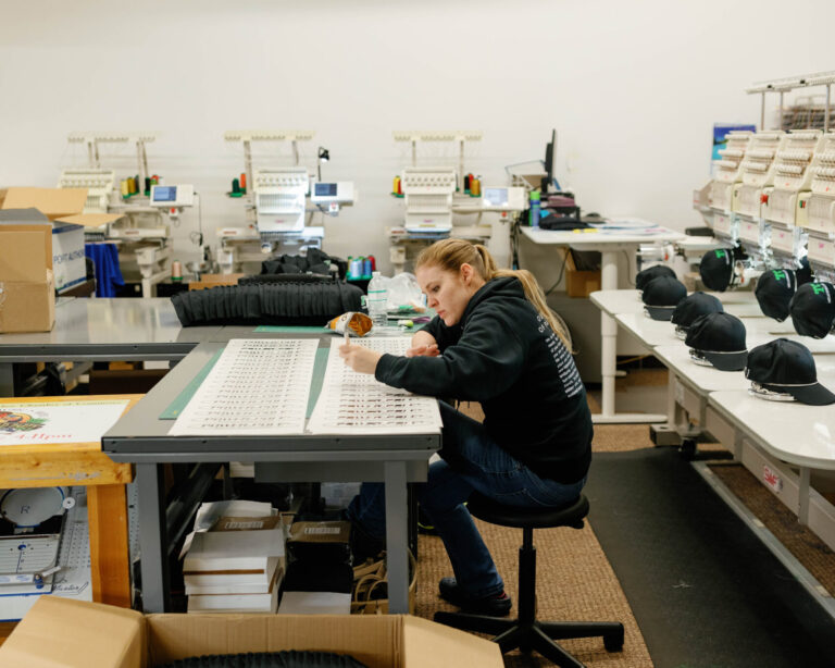 An IDX employee working on a design while a hat embroider machine works on hats behind her.