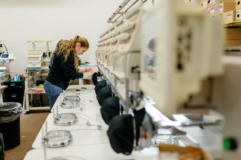 An IDX employee working on quality checking a hat that is getting embroidered by a machine.