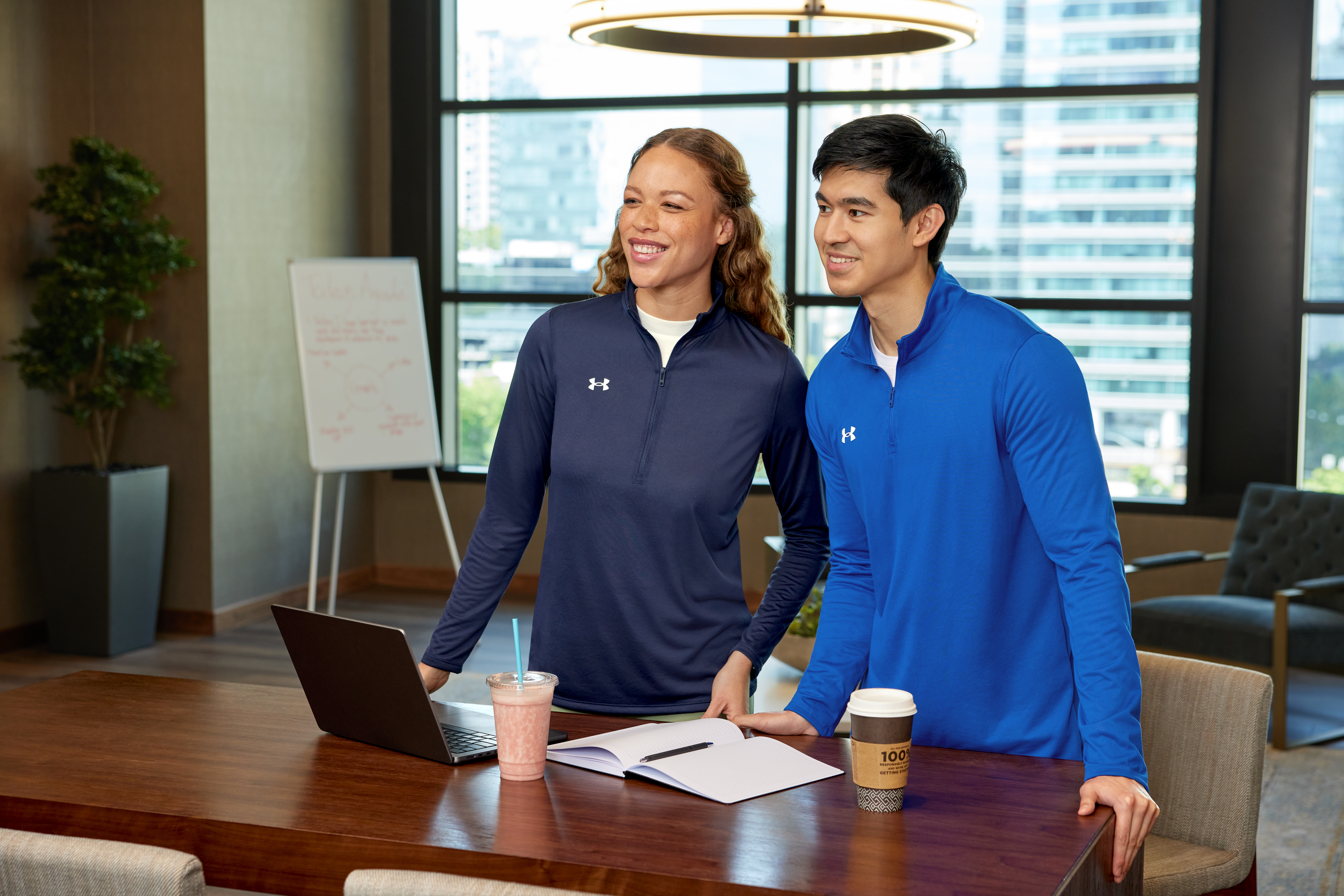 A woman and man in front of a desk at an event wearing under armor quarter zips in royal blue and navy smiling.