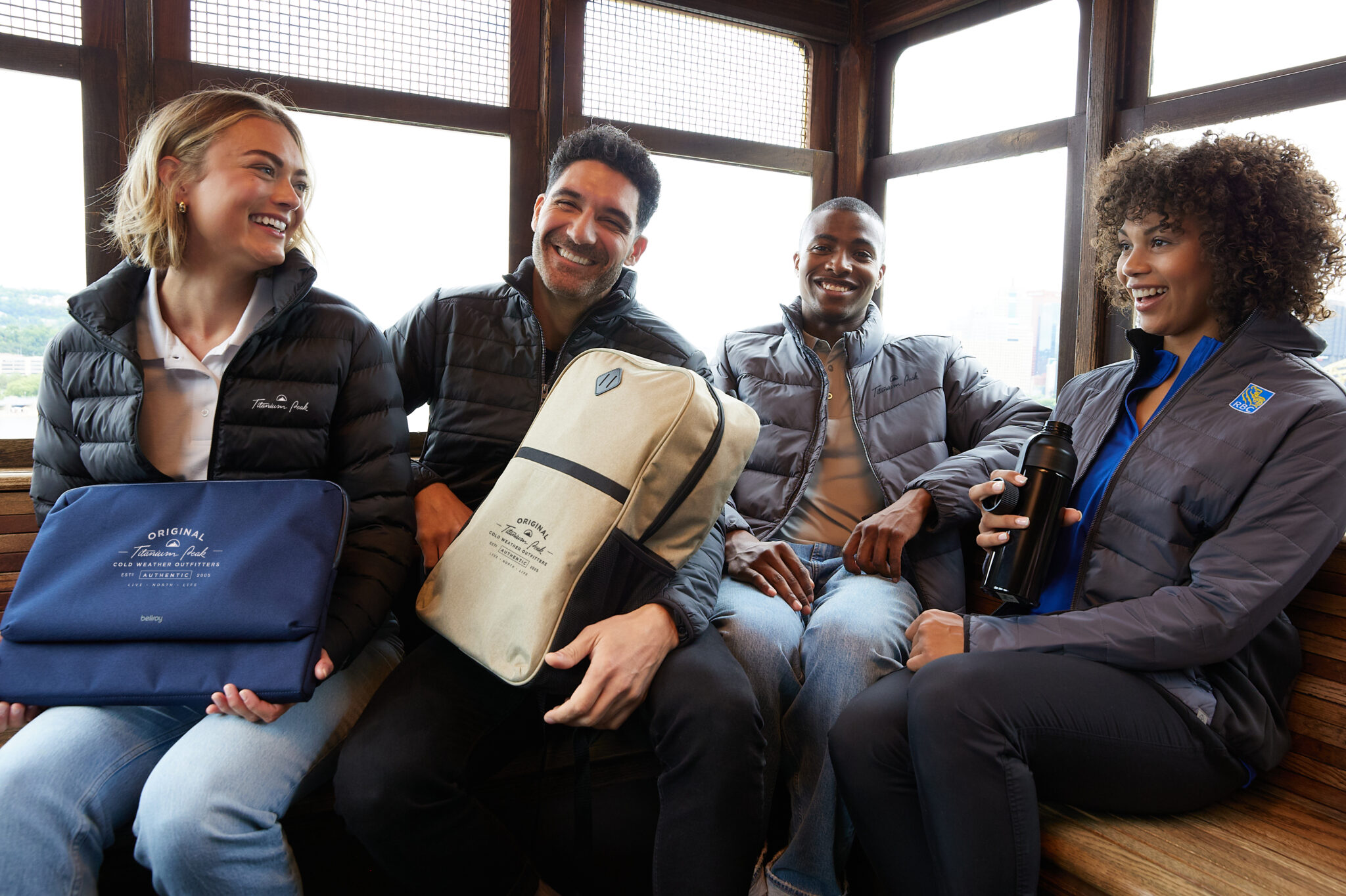 A group of different people sitting next to each other smiling and wearing jackets and backpacks. One woman is holding a black water bottle.