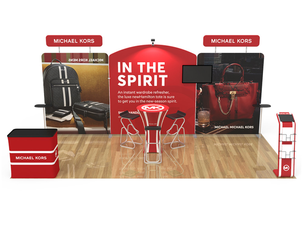 A 10x10 tradeshow banner in red and white with a picture that has a backpack and laptop bag, a purse, and a statement In The Spirit. A high top table and three chairs are in front of the banner.