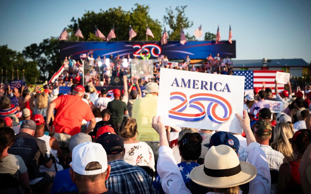 Large crowd at an America 250 public event holding branded signage and flags