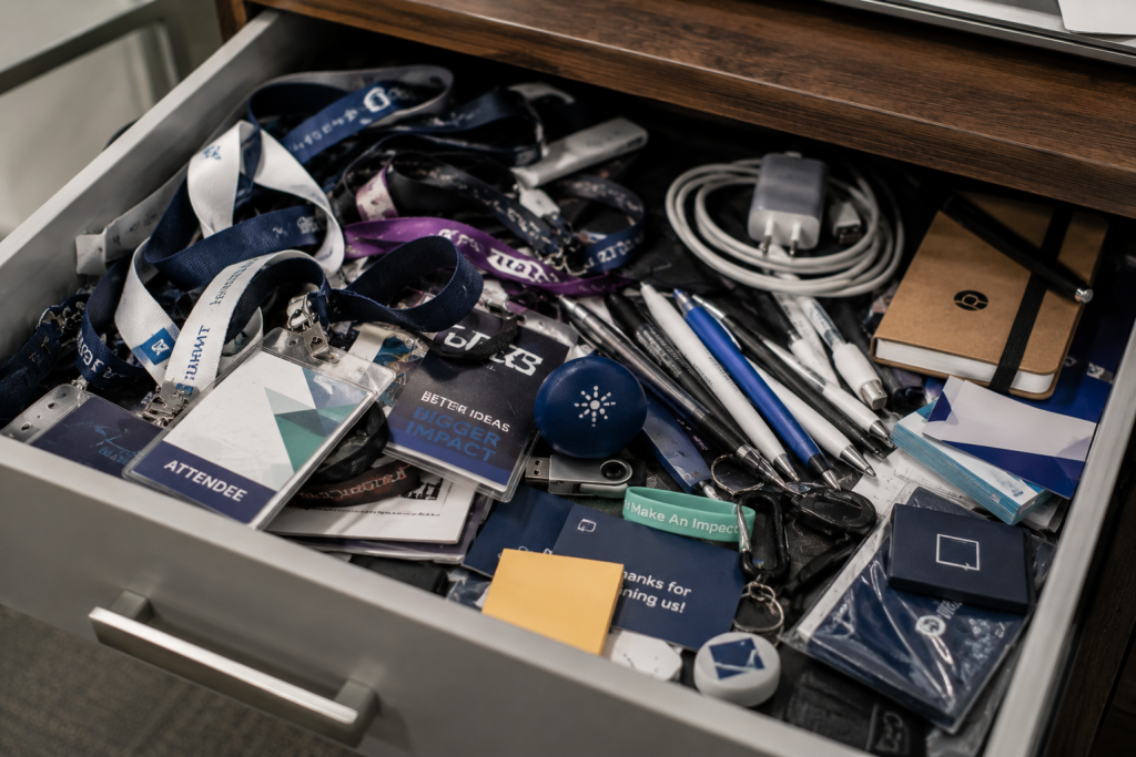 Open desk drawer overflowing with trade show giveaways including lanyards, badge holders, pens, USB drives, and branded cards