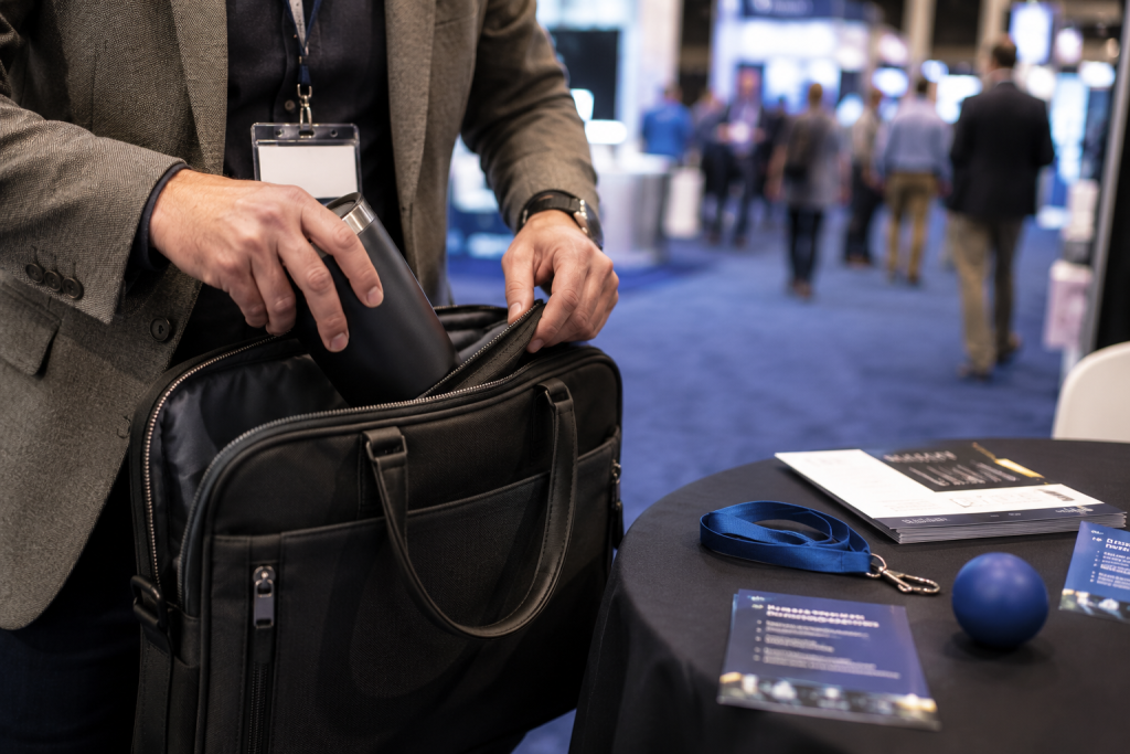 A trade show attendee placing a branded tumbler into their bag at a booth, with lanyards, brochures, and a stress ball visible on the table nearby.