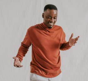 Smiling black man, wearing orange long sleeve with beige pants.