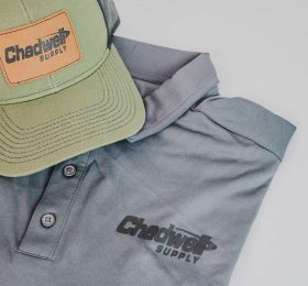 A light green hat with black mesh backing and a brown patch on the front that says Chadwell Supply on top of a gray collard polo shirt with the name logo name.